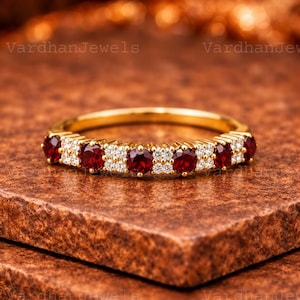 Könnte beinhalten: Ein Goldring mit einer Reihe abwechselnder runder roter Edelsteine und kleiner klarer Steine. Das Band hat eine polierte Oberfläche und der Text VardhanJewels ist im Hintergrund sichtbar. Das Design zeigt ein wiederkehrendes Muster aus roten und weißen Steinen.