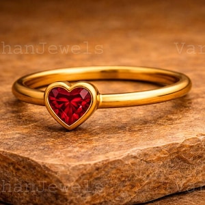 Può includere: Un anello d'oro con una pietra preziosa rossa a forma di cuore. L'anello è posto su una superficie marrone strutturata. La fascia dell'anello è sottile e lucida, e la gemma è di un colore rosso vibrante.