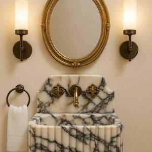 Könnte beinhalten: Luxuriöses Badezimmer-Waschtisch-Set mit Marmorwaschbecken mit Messingarmaturen, einem goldgerahmten ovalen Spiegel und passenden Wandleuchten. Ein weißes Handtuch hängt an einem Messingring. Das Waschbecken hat ein geripptes Design.