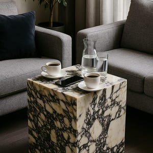 May include: A marble-effect cube table with two white coffee cups and saucers, a smartphone, a glass carafe, and a glass of water. The table is between two grey armchairs. A potted plant is in the background.