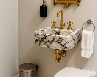 May include: A marble sink with gold fixtures, including a faucet and handles, is mounted on a wall. A gold-framed mirror hangs above the sink. A black soap dispenser and a stainless steel trash can are also visible.