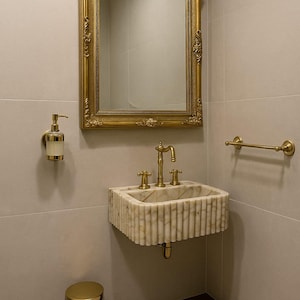 May include: A luxurious bathroom interior featuring a gold-framed mirror, a marble sink with gold fixtures, and a matching gold trash can. The walls are a neutral color, and a small rug is on the floor.
