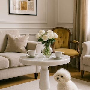 May include: A living room scene with a white marble table holding two teacups and a vase of white roses. A small white dog sits on the floor. A beige sofa and a yellow chair are in the background.