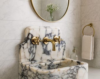 May include: A marble bathroom sink with gold fixtures. The sink has a rectangular basin and a decorative backsplash. A round gold mirror hangs above the sink, and a gold towel ring is on the wall. The sink is made of white marble with black veining.