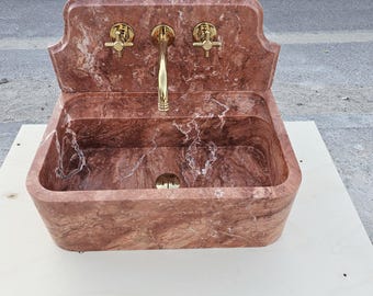 May include: A red marble sink with gold taps. The sink is rectangular with a decorative backsplash and a drain. The marble has white veining. The taps include a central spout and two separate handles.