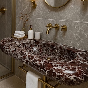 Può includere: Un lussuoso lavabo da bagno in marmo rosso con venature bianche. Il lavabo ha un design curvo ed è montato su una struttura in metallo color oro. Un asciugamano bianco è appeso a un portasciugamani dorato sotto il lavabo. Il bagno presenta rubinetteria e applique dorate.