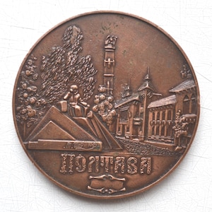 May include: A circular, bronze-toned medal with a detailed relief depicting a cityscape with buildings, trees, and a seated figure. The medal has text at the bottom and a blank space for engraving.