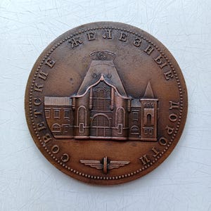 Puede incluir: Una moneda conmemorativa redonda de color bronce, con un diseño arquitectónico detallado de un edificio con una torre central y ventanas arqueadas. La moneda tiene texto cirílico alrededor del borde y un emblema estilizado en la parte inferior.