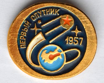 Puede incluir: Pin dorado con diseño azul y blanco. Presenta el texto "ПЕРВЫЙ СПУТНИК" en fuente curva, junto con una estrella roja y el número "1957". El diseño incluye una representación estilizada de un satélite y la Tierra.