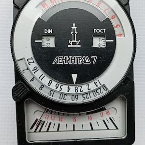 May include: A black Leningrad 7 light meter featuring a circular dial with white numerical scales for aperture and shutter speed settings. The device includes a central display with Cyrillic text, DIN and GOST markings, and a bottom window with a needle.