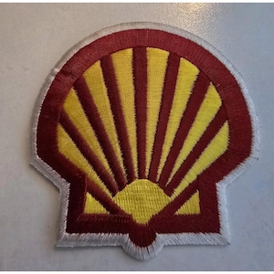 Op de afbeelding: Een geborduurde patch in de vorm van een sint-jakobsschelp met een felgeel centrum met stralende lijnen, omringd door een donkerrode rand en afgewerkt met een witte gestikte rand. Het ontwerp is symmetrisch en toont een klassiek waaierpatroon.