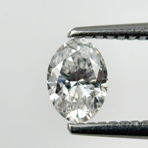 Puede incluir: Un diamante claro con talla marquesa, sostenido por unas pinzas plateadas. Las facetas del diamante reflejan la luz. El fondo es blanco, resaltando la claridad y forma de la gema.