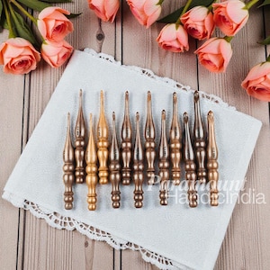 Puede incluir: Un conjunto de ganchillos de madera en varios tamaños y tonos marrones y dorados, dispuestos sobre un paño blanco con un borde decorativo. Los ganchillos se muestran con rosas rosas en el fondo, creando una imagen artesanal.