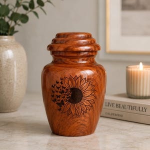 May include: A polished wooden cremation urn featuring a dark laser-engraved design of a sunflower on the front. The sunflower petals transition into a cluster of small butterflies. The urn has a rounded body and a tiered, removable lid.