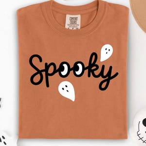 K&ouml;nnte beinhalten: Ein orangefarbenes T-Shirt mit dem Wort "Spooky" in schwarzer Schrift und wei&szlig;en Geistergrafiken. Das Wort "Yam" ist in schwarzer Schrift unten aufgedruckt. Das Shirt ist von der Marke Comfort Colors.