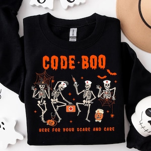 May include: Black sweatshirt with the text "CODE BOO" in orange. The design features skeleton nurses and a pumpkin, with the phrase "HERE FOR YOUR SCARE AND CARE" below. Halloween-themed apparel.