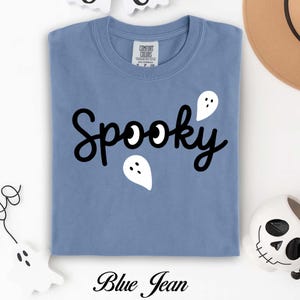 K&ouml;nnte beinhalten: Ein blaues Jeans-T-Shirt mit dem Wort "Spooky" in schwarzer Schrift und wei&szlig;en Geistergrafiken. Das Shirt hat einen Rundhalsausschnitt und kurze &Auml;rmel. Der Text "Blue Jean" ist unten auf dem Shirt aufgedruckt.