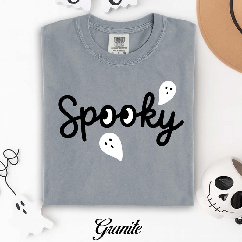 K&ouml;nnte beinhalten: Ein gefaltetes graues T-Shirt mit dem Wort "Spooky" in schwarzer Schrift, mit wei&szlig;en Geistergrafiken. Das Wort "Granite" ist in schwarzer Schrift unten aufgedruckt. Das Shirt ist von der Marke Comfort Colors.