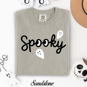 K&ouml;nnte beinhalten: Sandfarbenes T-Shirt mit dem Wort "Spooky" in schwarzer Schrift, begleitet von wei&szlig;en Geistergrafiken. Das Shirt ist von der Marke Comfort Colors. Zus&auml;tzliche Dekorationen zum Thema Halloween sind sichtbar.