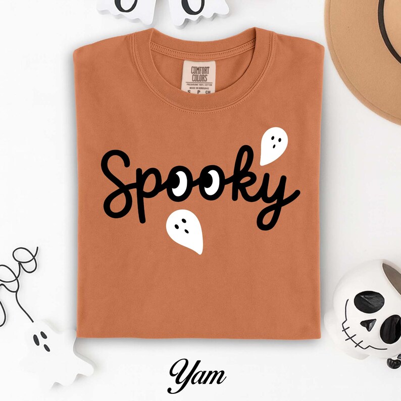 K&ouml;nnte beinhalten: Ein orangefarbenes T-Shirt mit dem Wort "Spooky" in schwarzer Schrift und wei&szlig;en Geistergrafiken. Das Wort "Yam" ist in schwarzer Schrift unten aufgedruckt. Das Shirt ist von der Marke Comfort Colors.
