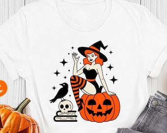 May include: White t-shirt featuring a Halloween-themed graphic. The design includes a witch sitting on a jack-o'-lantern, a raven, and a skull on books. The shirt is paired with gray jeans and surrounded by pumpkins and a candle.