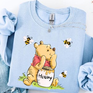 May include: A light blue sweatshirt featuring a cartoon illustration of Winnie the Pooh with a honey pot labeled "Hunny" and bees. The sweatshirt is paired with light wash denim jeans.