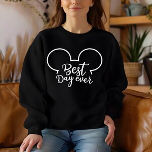 May include: Black sweatshirt with white graphic of Mickey Mouse ears and the text "Best Day ever". The sweatshirt is a crew neck style and appears to be made of a soft material. The design is simple and clean.