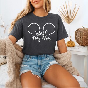 May include: Dark gray t-shirt with a white Mickey Mouse head outline and the script "Best Day Ever." The shirt is short-sleeved and made of a soft material. The model is wearing blue jean shorts and a beige sweater.