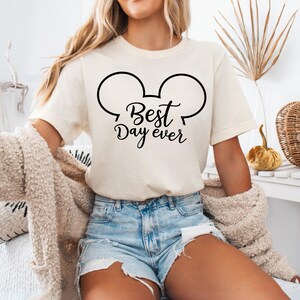 May include: Cream-colored t-shirt with a black graphic of Mickey Mouse ears and the words "Best Day Ever." The shirt is short-sleeved and made of a soft material. The shirt is paired with denim shorts.