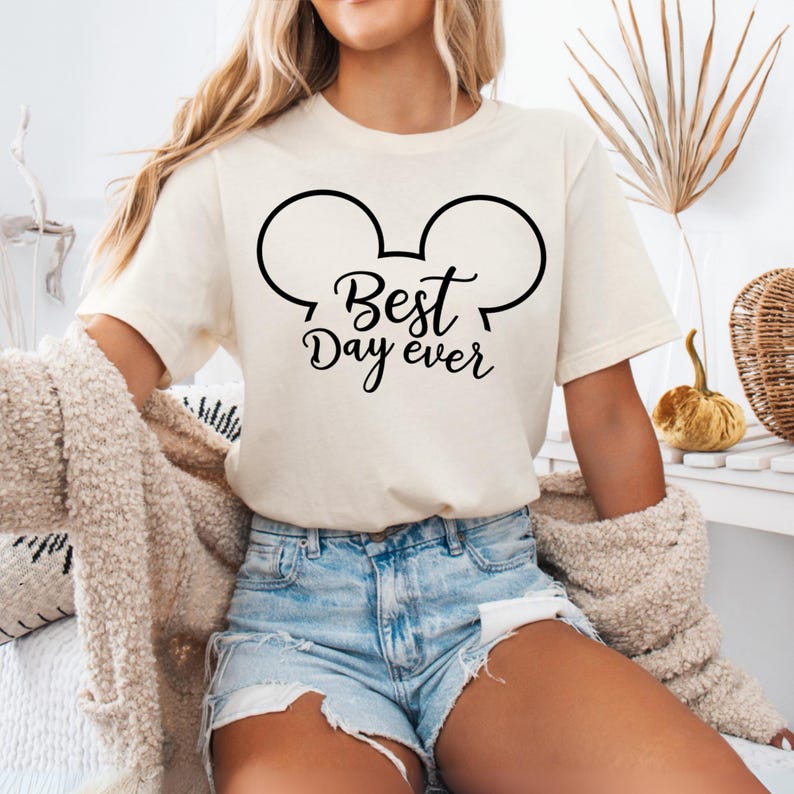 May include: Cream-colored t-shirt with a black graphic of Mickey Mouse ears and the words "Best Day Ever." The shirt is short-sleeved and made of a soft material. The shirt is paired with denim shorts.