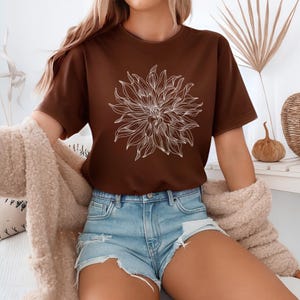 May include: A brown t-shirt with a white floral graphic. The short-sleeved top is paired with denim shorts. The image suggests a casual, everyday style. The shirt is made of a soft material.