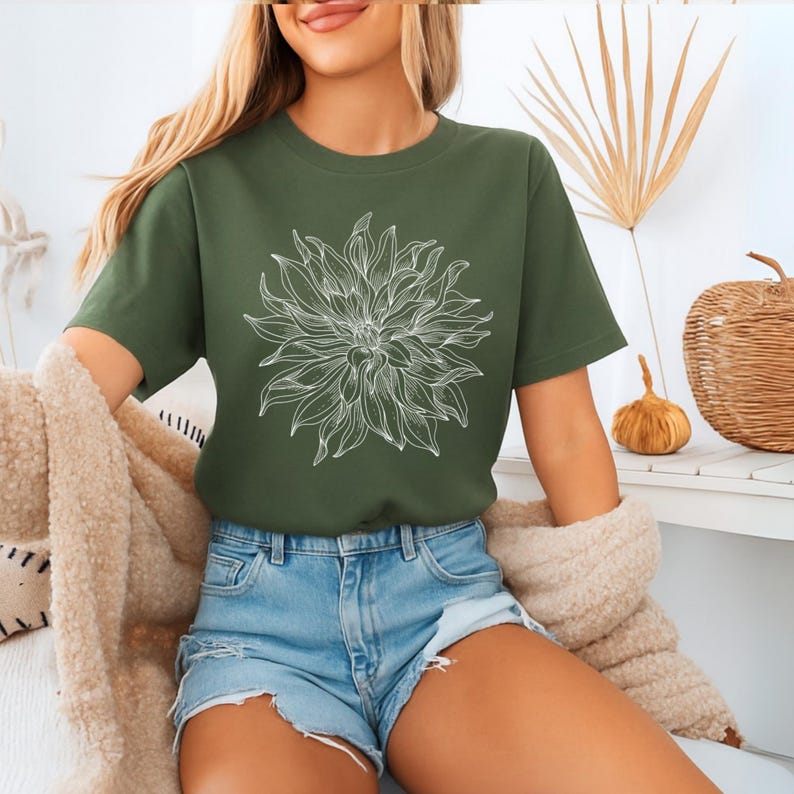 May include: An olive green t-shirt with a white floral design. The short-sleeved top is paired with denim shorts. The image suggests a casual, comfortable style, ideal for everyday wear. The shirt is made of a soft material.