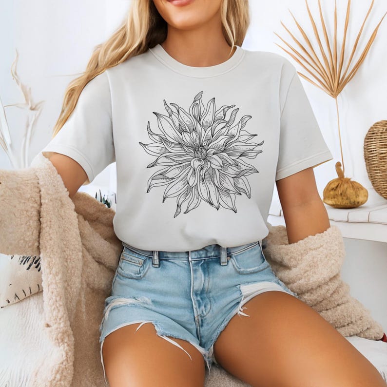 May include: A cream-coloured t-shirt with a detailed black and white floral design. The graphic is a large, stylized dahlia. The shirt has short sleeves and a crew neck. The model is wearing denim shorts and a beige jacket.