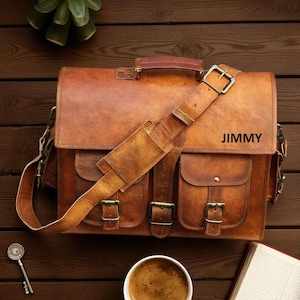May include: A brown leather satchel with a shoulder strap and multiple pockets. The bag has a handle and metal buckles. The word "JIMMY" is printed on the front. The bag is sitting on a wooden surface.