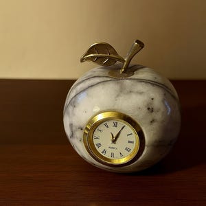 May include: A marble apple-shaped clock with a gold-tone clock face and stem and leaf detail. The clock face has Roman numerals. The apple has a white and gray marbled pattern. A decorative accent piece for a desk or shelf.