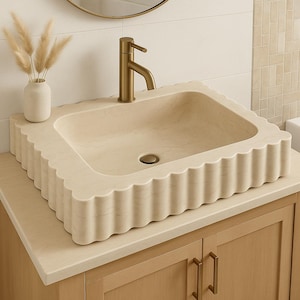 Può includere: Un lavabo rettangolare beige con bordo smerlato. Un rubinetto dorato è montato sul lavabo. Il lavabo è appoggiato su un mobile bagno in legno chiaro con maniglie dorate. Un piccolo vaso con piante secche è appoggiato sul bancone.