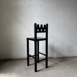 Pu&ograve; includere: Uno sgabello da bar in legno nero con un design dello schienale unico. Lo schienale presenta tre fori circolari e una forma a corona. Lo sgabello ha un sedile quadrato e quattro gambe robuste, con supporti orizzontali. Lo sgabello &egrave; nero.