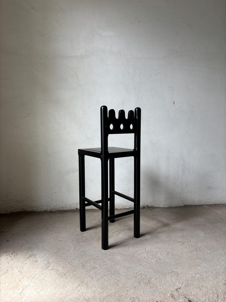 Pu&ograve; includere: Uno sgabello da bar in legno nero con un design dello schienale unico. Lo schienale presenta tre fori circolari e una forma a corona. Lo sgabello ha un sedile quadrato e quattro gambe robuste, con supporti orizzontali. Lo sgabello &egrave; nero.