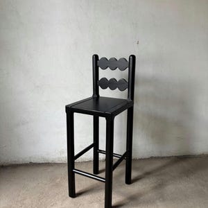 Op de afbeelding: Een zwarte houten barkruk met een vierkante zitting en een rugleuning met twee rijen geschulpte uitsparingen. De kruk heeft vier poten die met een voetensteun zijn verbonden. Het ontwerp is minimalistisch en modern. Afgewerkt in matzwart.