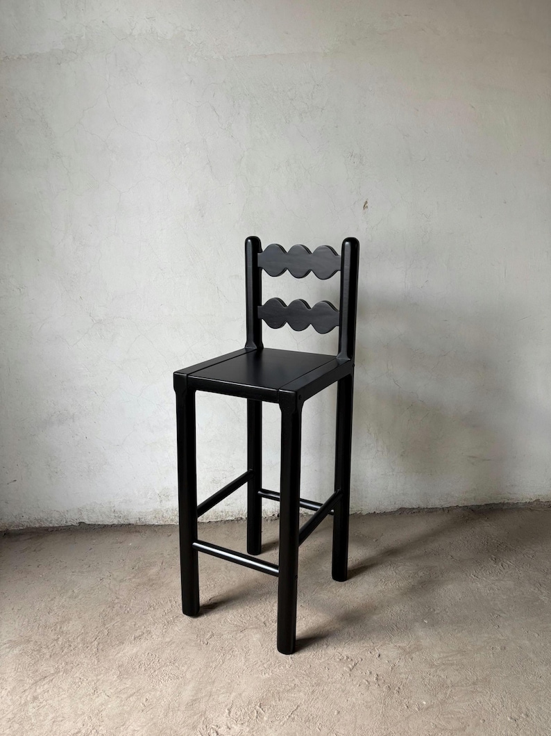 Op de afbeelding: Een zwarte houten barkruk met een vierkante zitting en een rugleuning met twee rijen geschulpte uitsparingen. De kruk heeft vier poten die met een voetensteun zijn verbonden. Het ontwerp is minimalistisch en modern. Afgewerkt in matzwart.