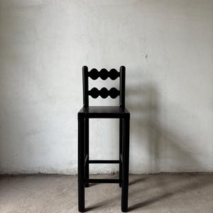 Op de afbeelding: Een zwarte houten barkruk met een decoratieve rugleuning met twee rijen van drie gebogen uitsparingen. De kruk heeft een vierkante zitting en vier rechte poten. Achtergrond: een effen, gebroken witte muur en een betonnen vloer.