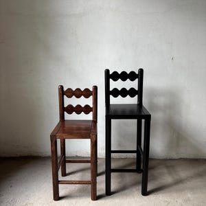 Op de afbeelding: Twee houten barkrukken. E&eacute;n is donkerbruin gebeitst, de andere is zwart geschilderd. Beide hebben een geschulpte rugleuning en een vierkante zitting. De krukken staan tegen een neutraal gekleurde muur.
