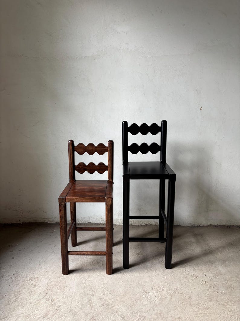 Op de afbeelding: Twee houten barkrukken. E&eacute;n is donkerbruin gebeitst, de andere is zwart geschilderd. Beide hebben een geschulpte rugleuning en een vierkante zitting. De krukken staan tegen een neutraal gekleurde muur.