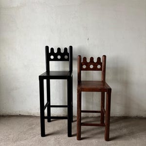 Pu&ograve; includere: Due sgabelli da bar in legno. Uno &egrave; verniciato di nero, l'altro &egrave; in tonalit&agrave; legno naturale. Entrambi hanno uno schienale decorativo con tre ritagli circolari e un bordo superiore smerlato. Gli sgabelli hanno un poggiapiedi e sono contro un muro bianco.