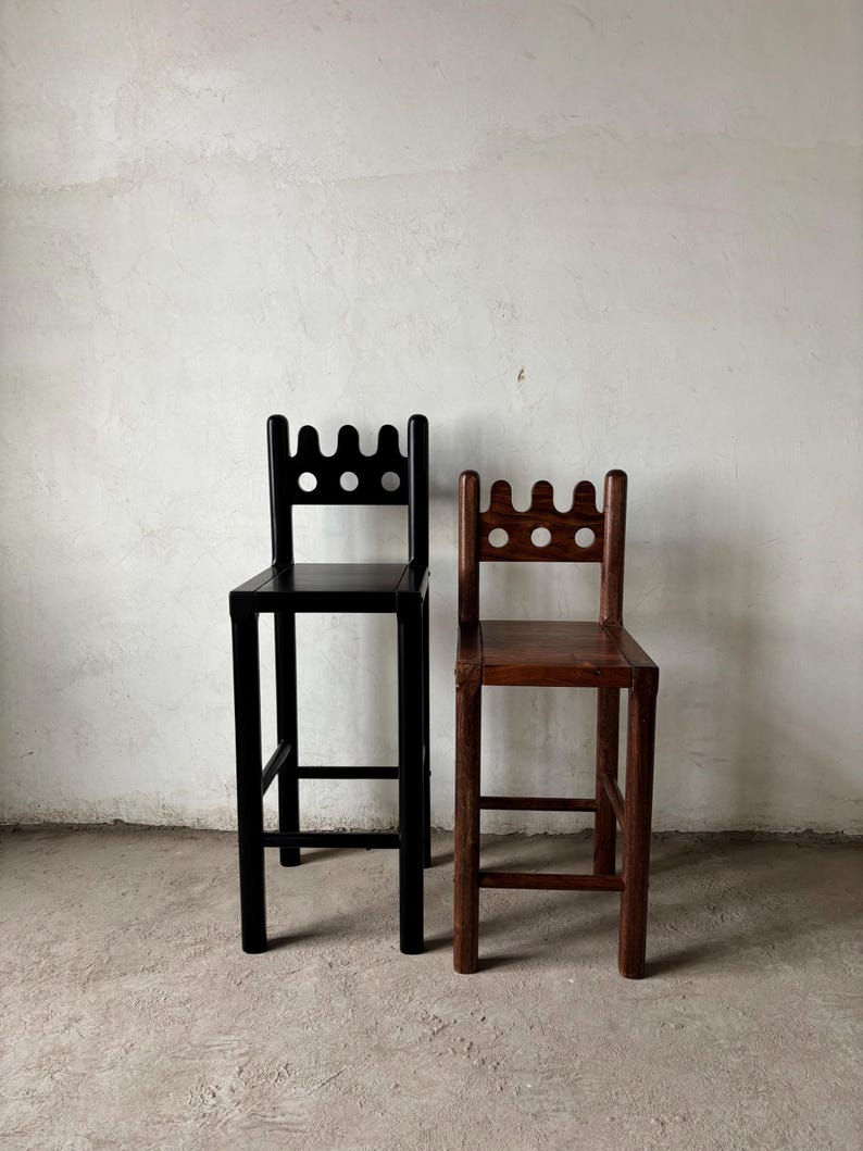 Pu&ograve; includere: Due sgabelli da bar in legno. Uno &egrave; verniciato di nero, l'altro &egrave; in tonalit&agrave; legno naturale. Entrambi hanno uno schienale decorativo con tre ritagli circolari e un bordo superiore smerlato. Gli sgabelli hanno un poggiapiedi e sono contro un muro bianco.