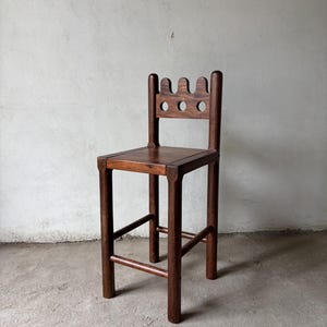Pu&ograve; includere: Sgabello da bar in legno marrone scuro con un design unico dello schienale. Lo schienale presenta tre fori circolari e un bordo superiore ondulato. Lo sgabello ha un sedile quadrato e gambe robuste collegate da supporti orizzontali.