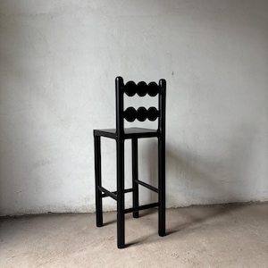 Op de afbeelding: Een zwarte houten barkruk met een uniek rugleuningontwerp. De rugleuning heeft een reeks cirkelvormige uitsparingen. De kruk heeft een vierkante zitting en vier rechte poten, wat een minimalistische esthetiek cre&euml;ert. Hoogte ongeveer 76 cm.