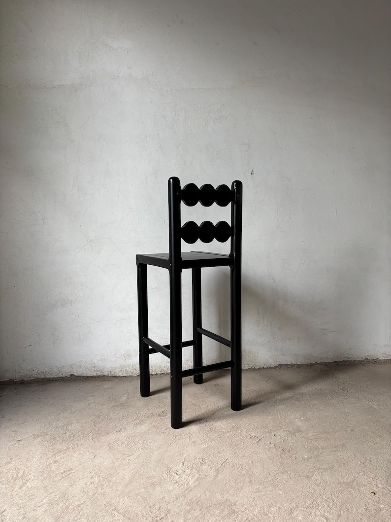Op de afbeelding: Een zwarte houten barkruk met een uniek rugleuningontwerp. De rugleuning heeft een reeks cirkelvormige uitsparingen. De kruk heeft een vierkante zitting en vier rechte poten, wat een minimalistische esthetiek cre&euml;ert. Hoogte ongeveer 76 cm.