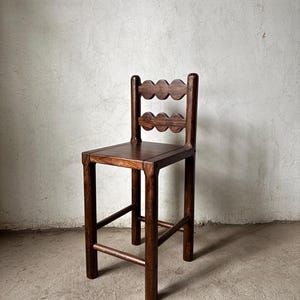 Op de afbeelding: Donkerbruine houten stoel met een vierkante zitting en een rugleuning met een geschulpte vormgeving. De stoel heeft vier poten en een voetensteun. De stoel is gemaakt van hout en heeft een eenvoudig, rustiek ontwerp.