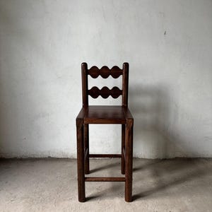 Op de afbeelding: Een hoge, donkerbruine houten barkruk met een geschulpte rugleuning. De kruk heeft een vierkante zitting en vier stevige poten die door een voetensteun zijn verbonden. De houtnerf is zichtbaar, wat hem een rustieke uitstraling geeft.